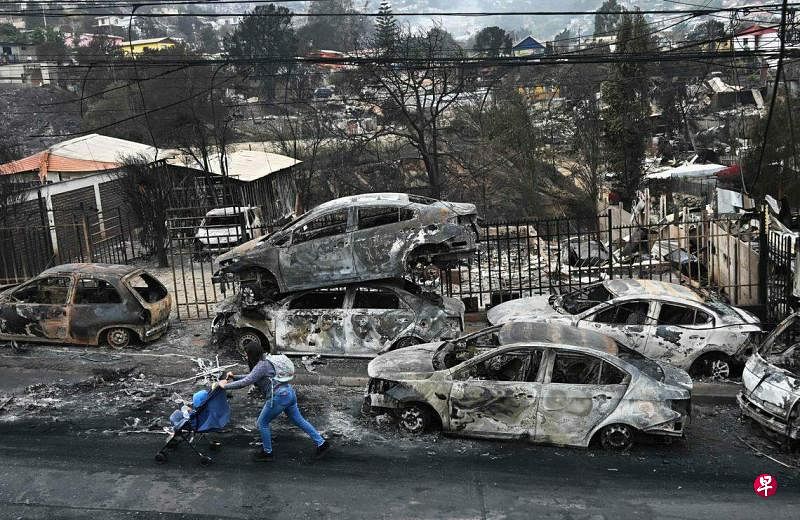 智利森林火灾灾情严重，图为一名妇女推着宝宝经过许多被大火烧毁的车辆。（法新社）