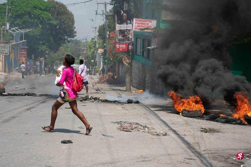 3月20日，海地首都太子港响起枪声，一名妇女抱着孩子在街上奔逃躲避，她身后的轮胎起火，冒出黑烟，街道满目狼藉。（法新社）